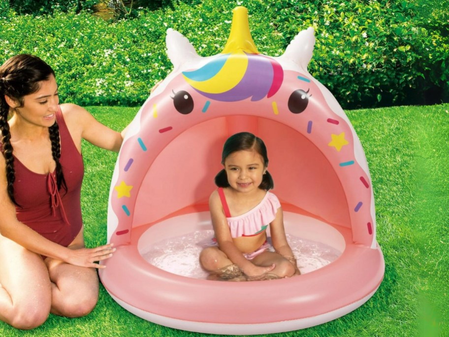little girl in a pink unicorn inflatable shade pool with mom sitting next to it