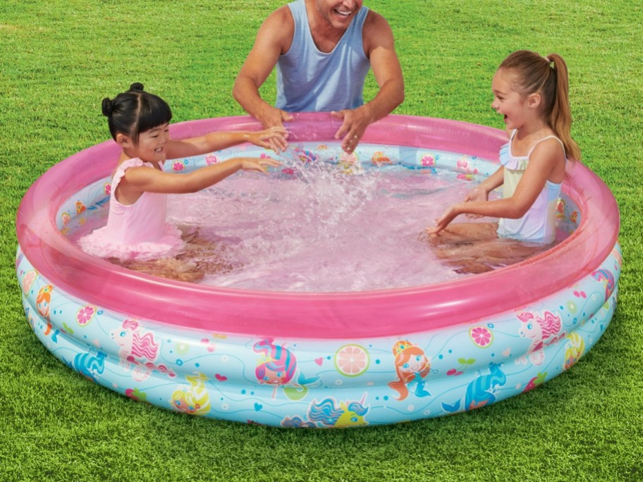little girls playing in an inflatable pool that is pink and blue with mermaids on it