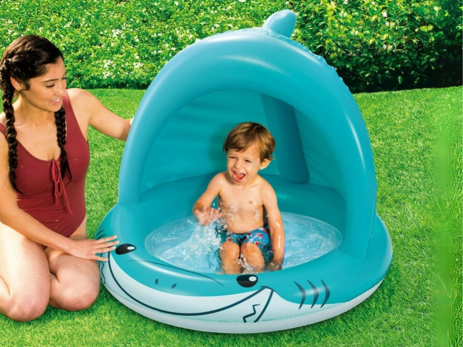 little boy in a blue shark inflatable shade pool with mom sitting next to it