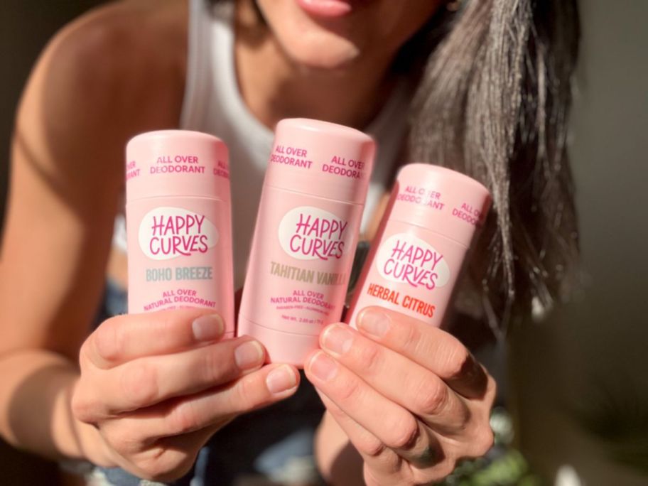 A woman holding 3 Happy Curves full body deodorant tubes