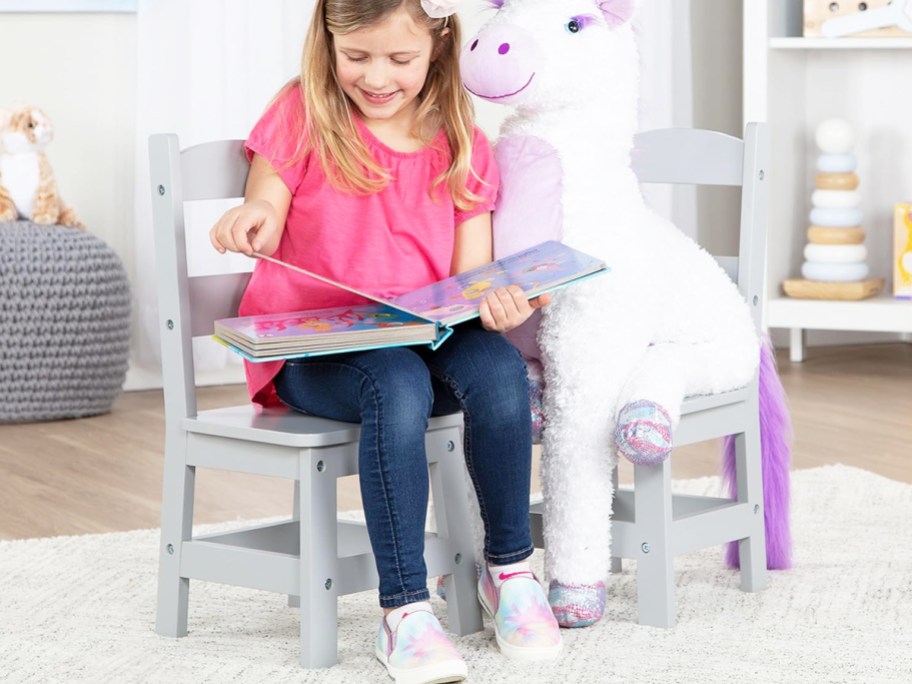 girl sitting on wooden chair reading a book next to a giant unicorn plush