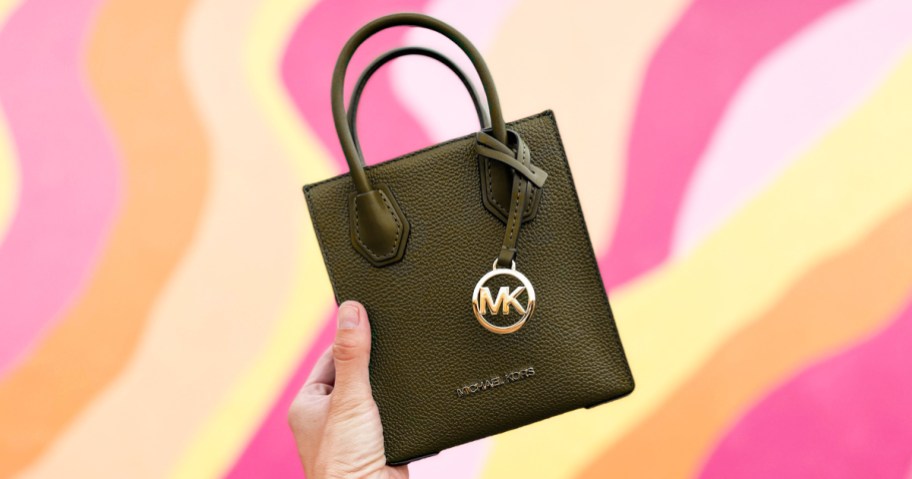 hand holding up a small Michael Kors bag against a pink, orange, and yellow wall