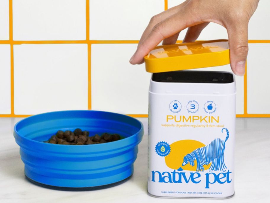 A bowl of dog food with a human hand opening a tin of native pet pumpkin next to it