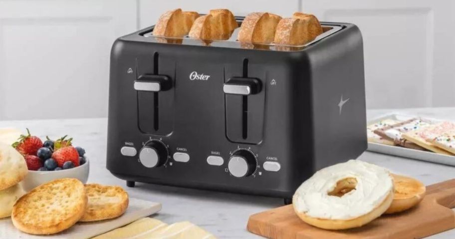 An Oster 4-slice toaster with 4 slices of toast in it surrounded by plates of toasted foods like bagels, toaster pastries and english muffins