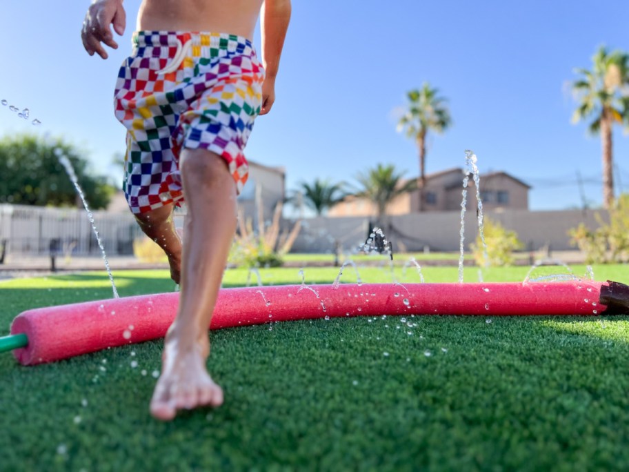 boy running through a diy pool noodle sprinkler, one of the easy dollar tree summer activities for kids