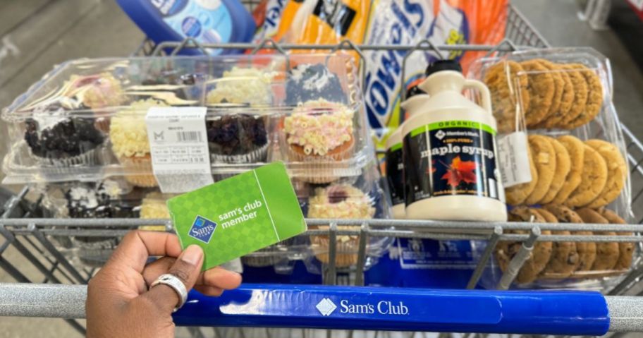 A sam's Club cart full of groceries with a hand holding a Sam's Club membership card