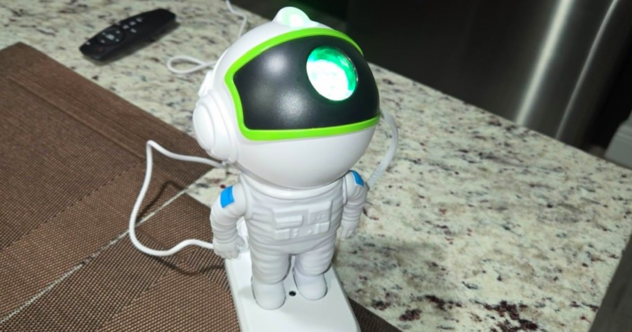An astronaut galaxy star projector with a glowing green light in its visor, sitting on a kitchen counter next to a remote control.