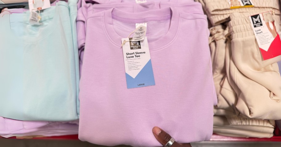 hand reaching for a light purple t-shirt, next to a blue t-shirt and tan pair of shorts on a display table at Sam's Club