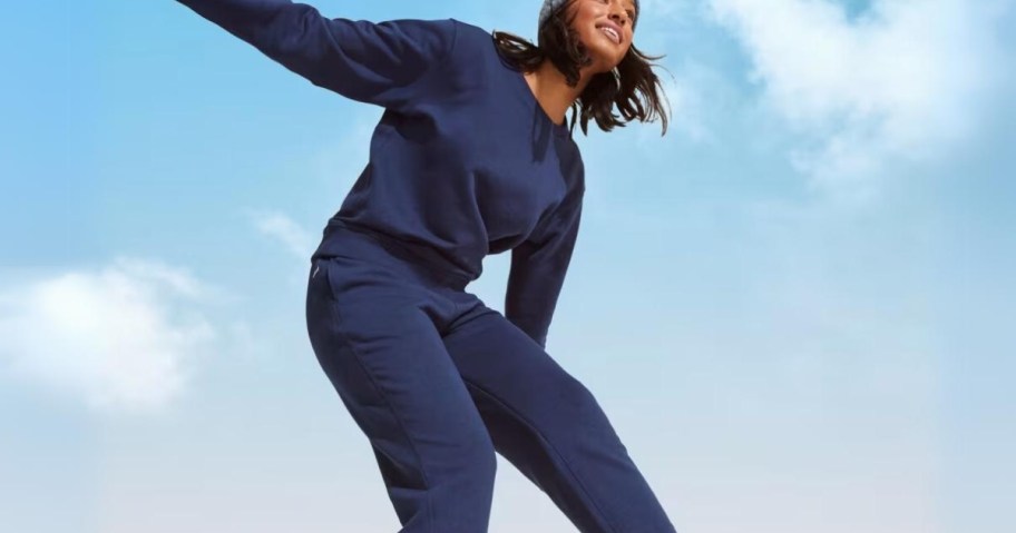 woman wearing navy blue sweatshirt and sweatpants with her arm out, blue sky behind her