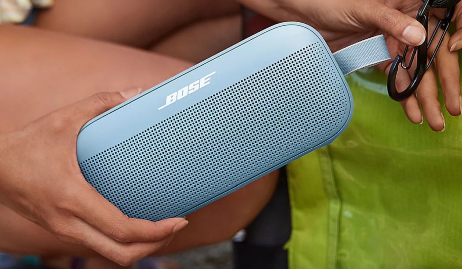 hands holding bluish grey bose speaker on carabiner