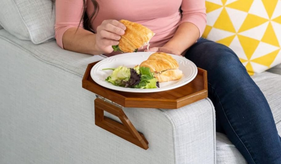 a woman eating a chicken salad sandwich with plate sitting on a brown wooden octagonal clip on couch arm table