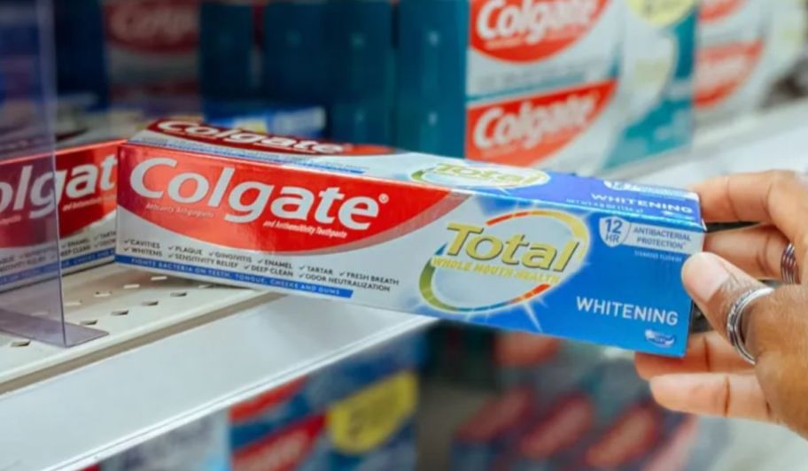 a womans hand grabbing a box of colgate total whitening toothpaste from a store shelf