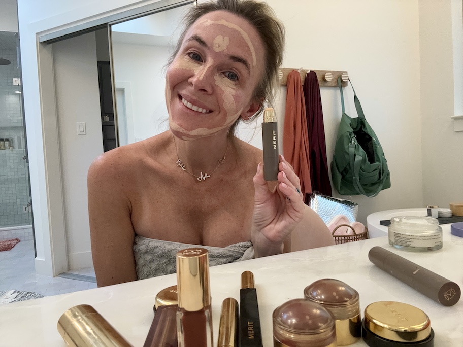 woman holding up merit makeup in towel sitting in bathroom