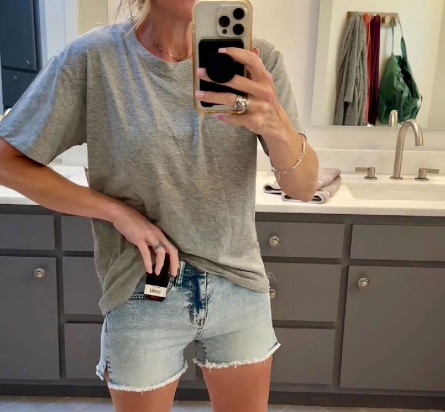 woman taking selfie in mirror wearing light wash shorts and tshirt