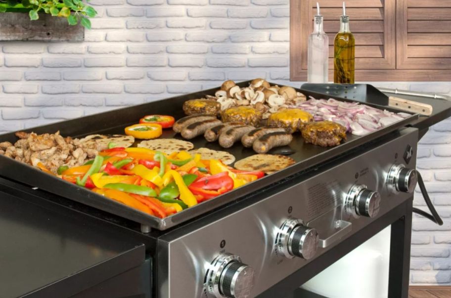 a large outdoor gas griddle with onions peppers, sausages, burgers and chicken cooking on its surface