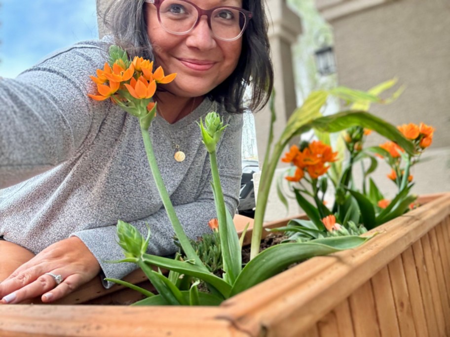 smiling woman leaning on a wooden planter filled with soil and orange and yellow flowers