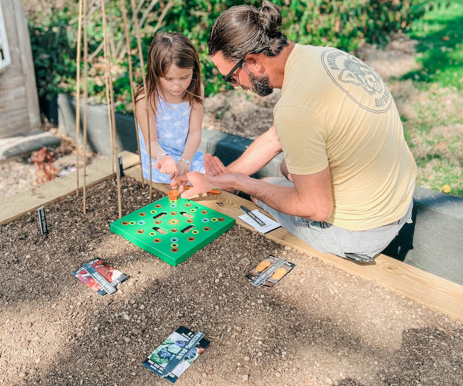 girl and man sitting on raised garden bed planting seeds in soil with tray