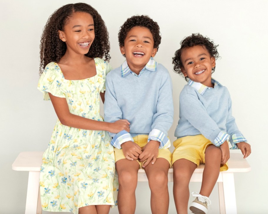 girl wearing yellow floral dress and two boys wearing blue long sleeve shirts sitting on a white bench