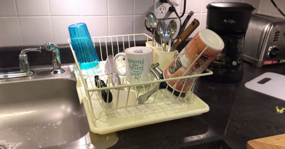 a white dish draining rack with dishes drying on it,on a green counter top next to a stainless steel sink