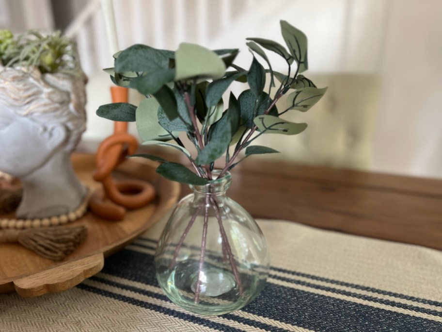 hand touching the leaves of a faux eucalyptus plant in a glass vase