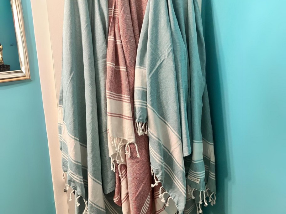 3 different color striped large beach towels with fringe hanging on hooks on a bathroom wall