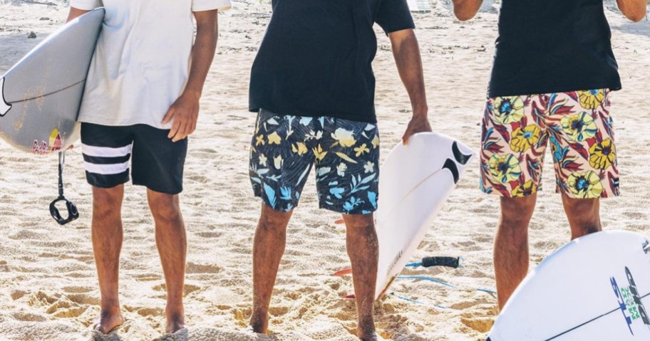 3 men on a beach wearing Hurley board shorts and t-shirts and holding surfboards