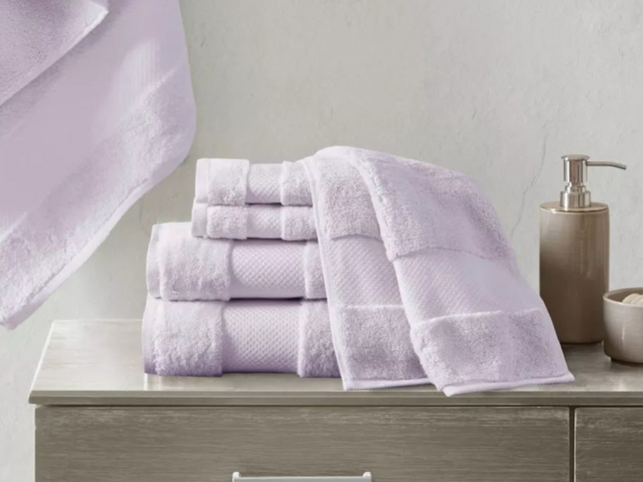 set of lavender color bath towels, hand towels and wash cloths on a bathroom counter, 1 hanging on the wall