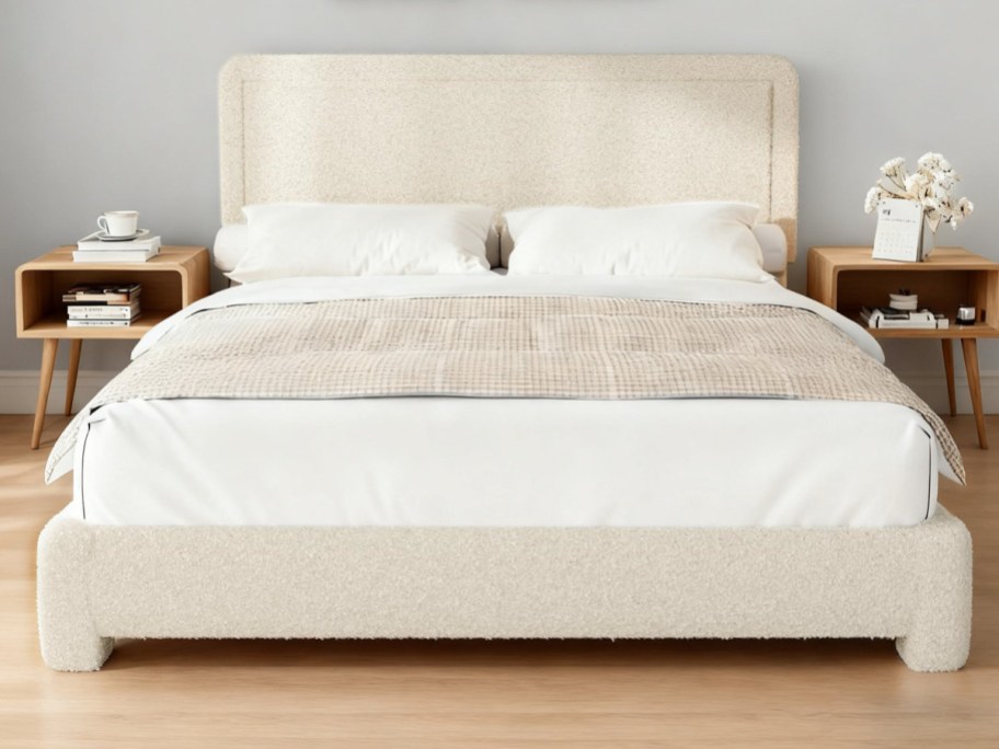 A cozy bedroom features a beige bed with a textured headboard, white pillows, and a light brown quilt. Two wooden bedside tables hold books, a cup, and flowers.