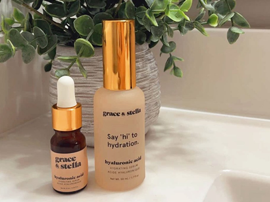 small and large bottles of Grace & Stella Hyaluronic Acid Serum on bathroom counter in front of plant