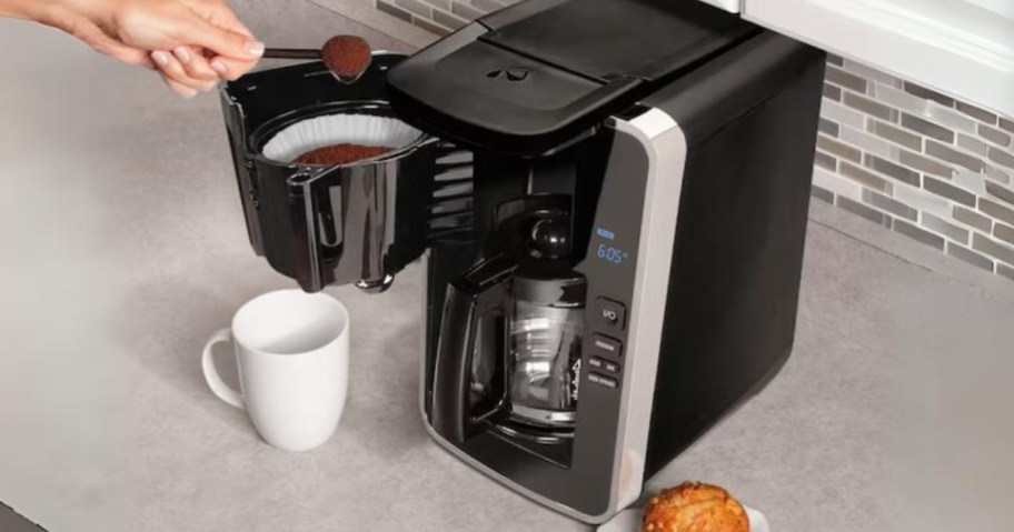 person putting ground coffee into a black and silver coffee maker sitting on a counter next to a mug and muffin