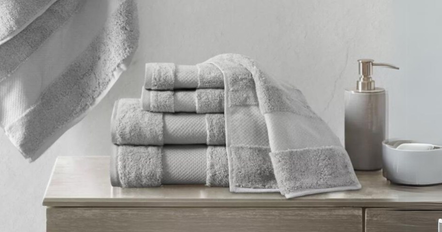 set of light grey color bath towels, hand towels and wash cloths on a bathroom counter, 1 hanging on the wall
