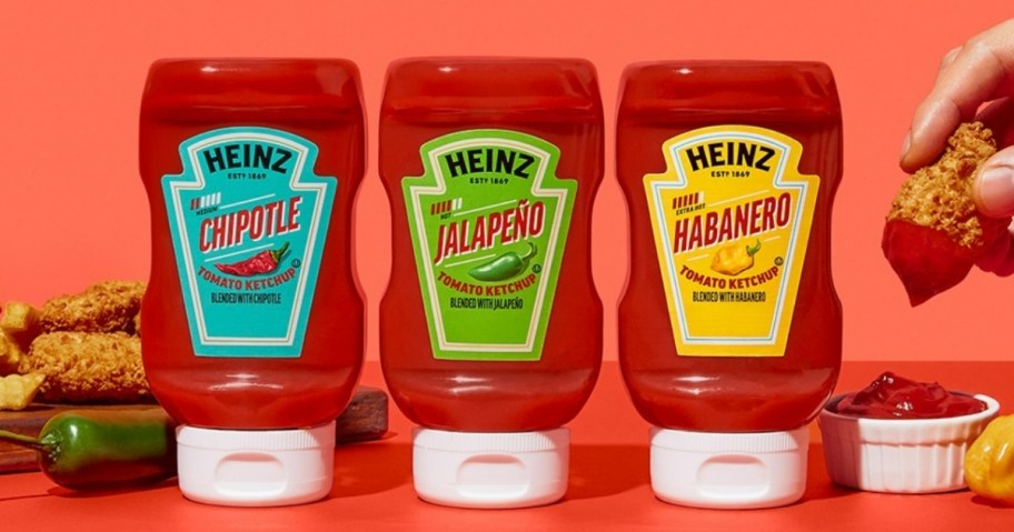 3 Heinz Spice Ketchup bottles next to a pepper and person's hand with chicken tender dipped in 1 of the flavors