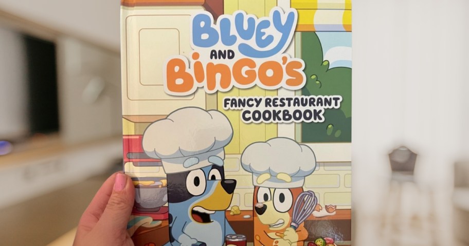 hand holding a Bluey and Bingo's Fancy Restaurant Cookbook with Bluey & Bingo on the cover wearing chefs hats