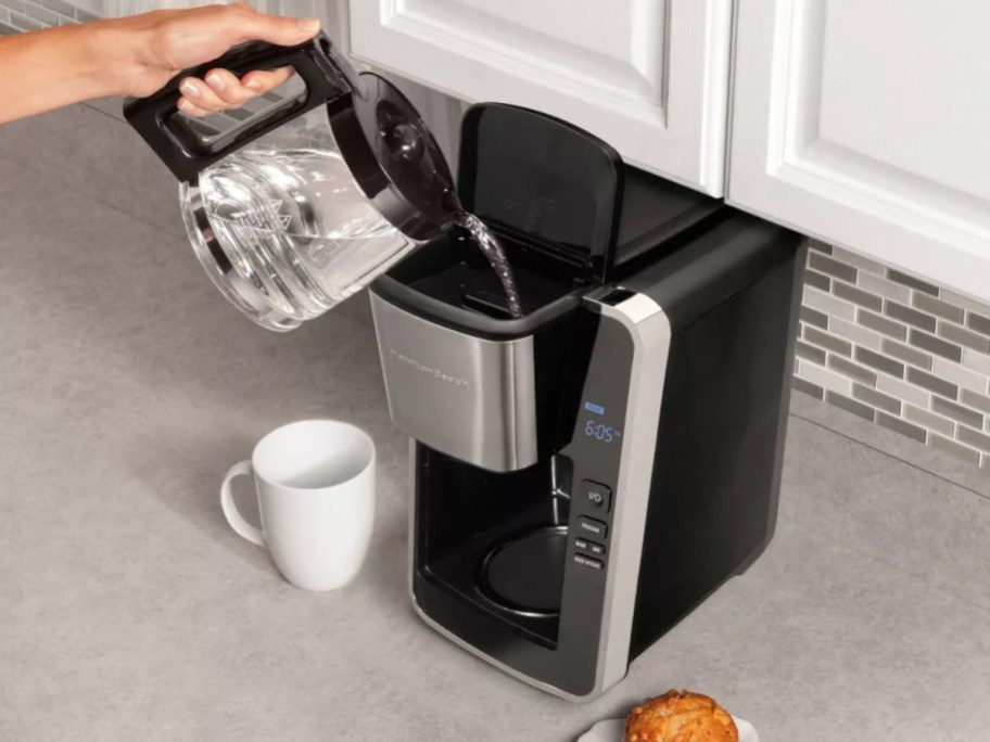 person pouring water into the top of a black and silver coffee maker sitting on a counter next to a mug and muffin