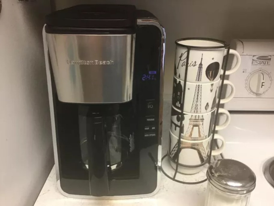 black and silver Hamilton Beach Coffee maker next to a set of 4 mugs and sugar dispenser on a kitchen counter