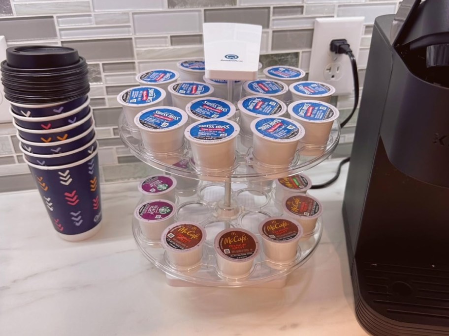 2-tier k-cup organizer with k-cups in it next to a Keurig machine and coffee cups on a counter
