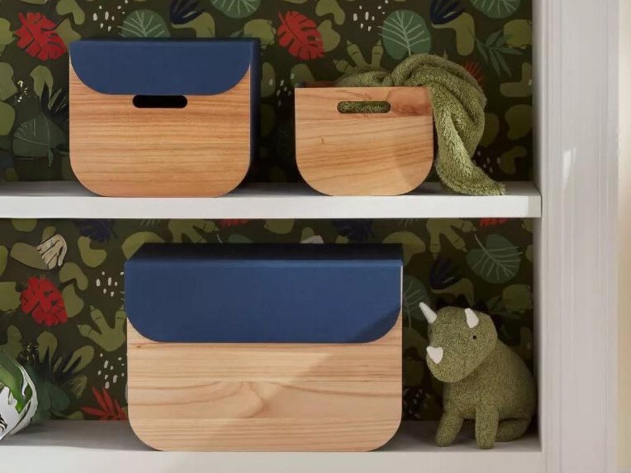 3 wooden storage bins with navy fabric lids in a bookcase with floral wallpaper on the back