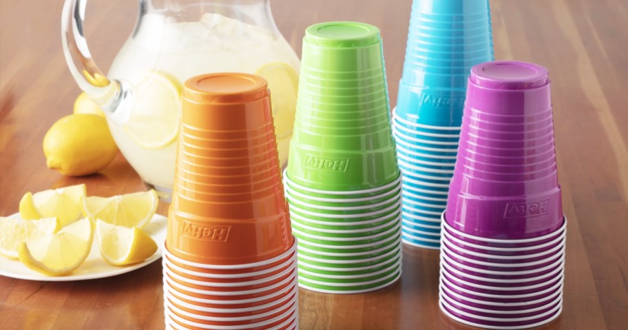 stacks of orange, green, blue, and purple disposable cups on table in front of pitcher of lemonade