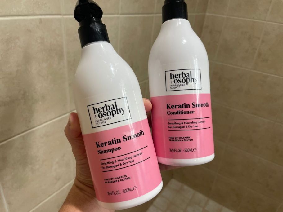 A hand holding a Herbalosophy Biotin + Collagen Shampoo and Conditioner in a shower