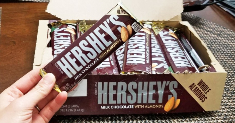 hand holding up a bar of Hershey's Milk Chocolate with Whole Almonds with large box of more bars behind them