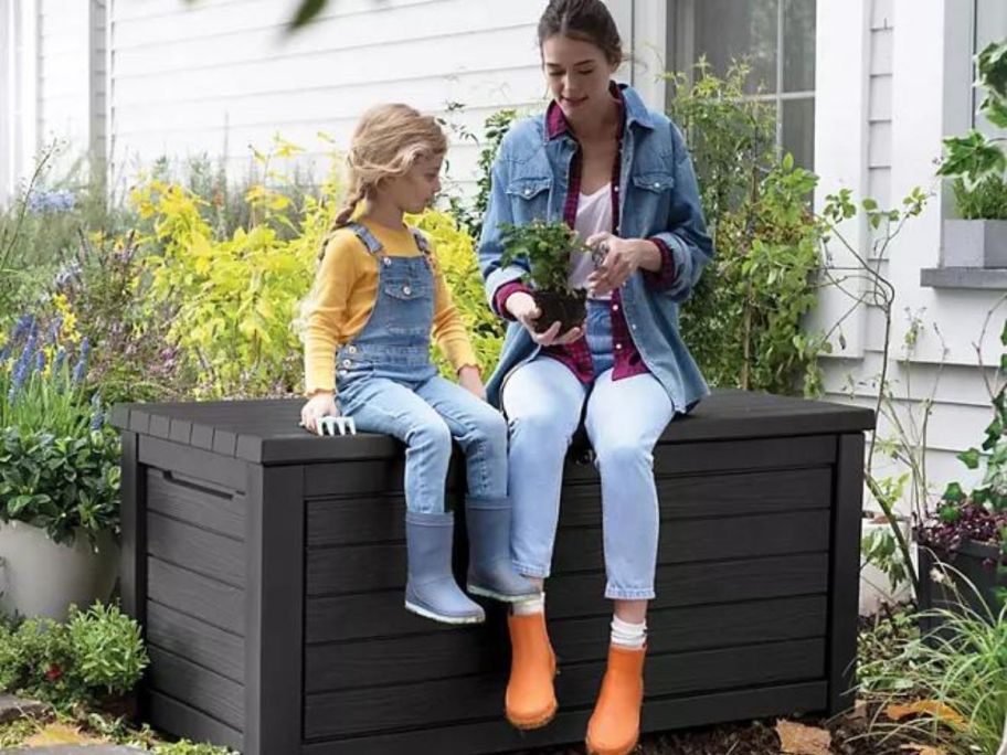 little girl and woman sitting on a Keter deck box looking at a plant