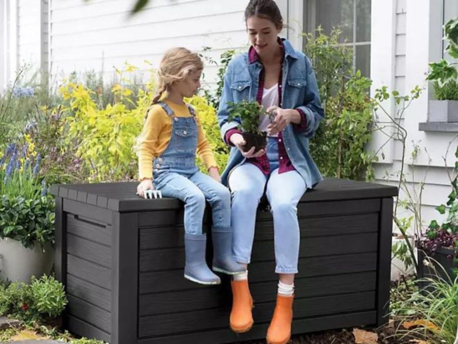 little girl and woman sitting on a Keter deck box looking at a plant