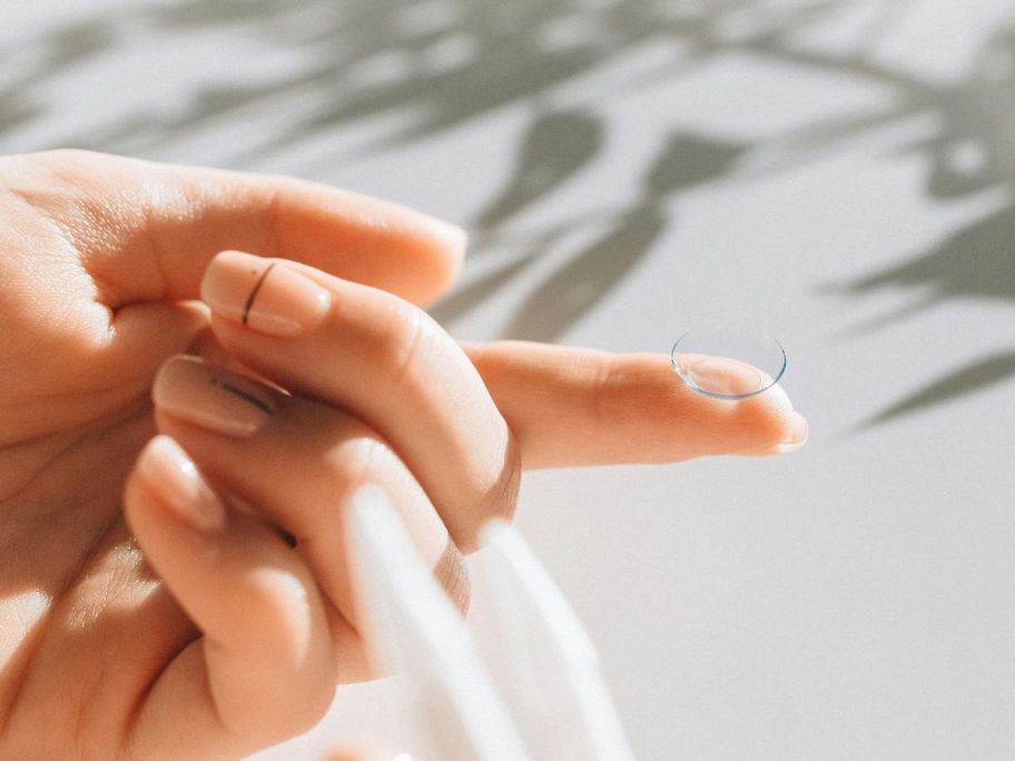 Hand with plastic tweezers in one hand and a contact lens on its extended index finger on the other hand