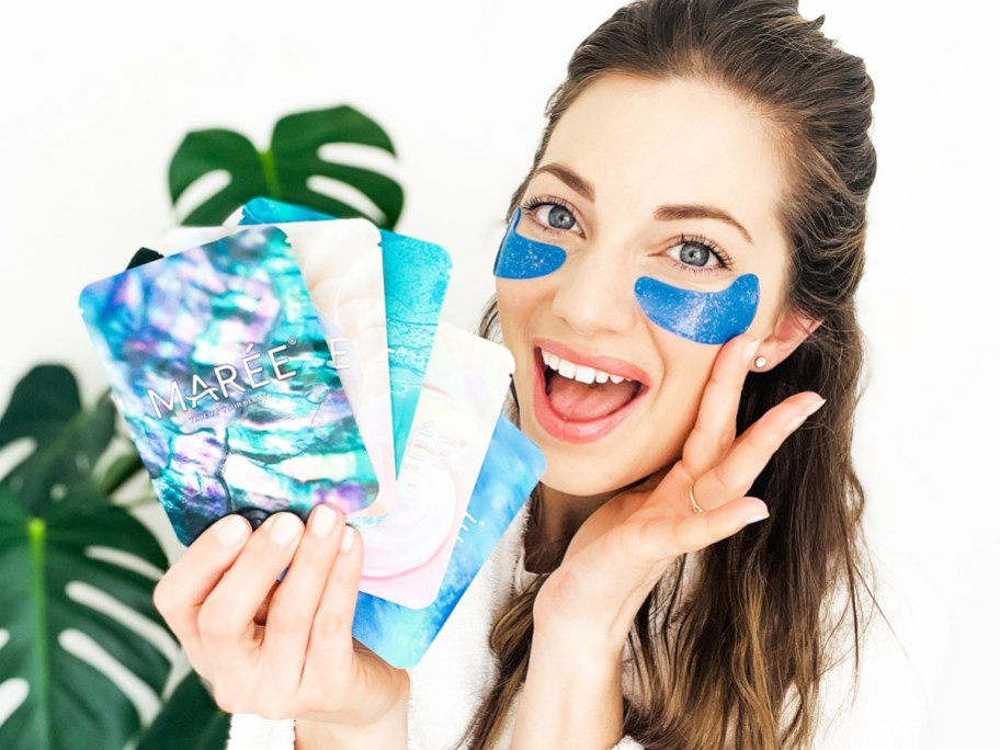 woman holding up multiple packs of eye gels while wearing a blue pair under her eyes