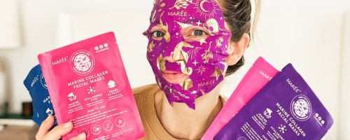 woman holding up multiple packs of sheet masks while wearing a pink and gold sheet mask on her face