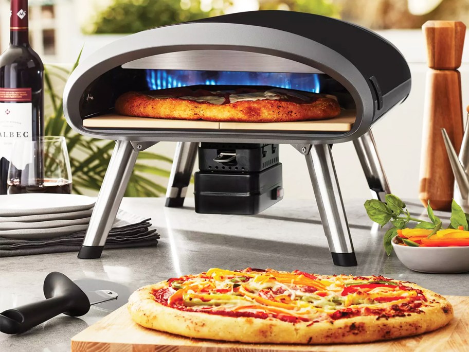 black tabletop pizza oven with pizza inside and one in front of it on a cutting board