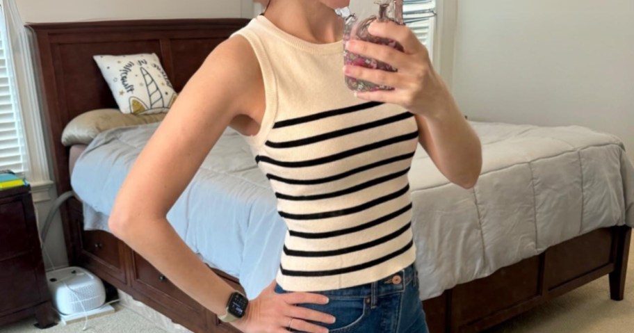 woman taking a picture of herself in a bedroom, she's wearing an off white and black stripe tank top and jeans