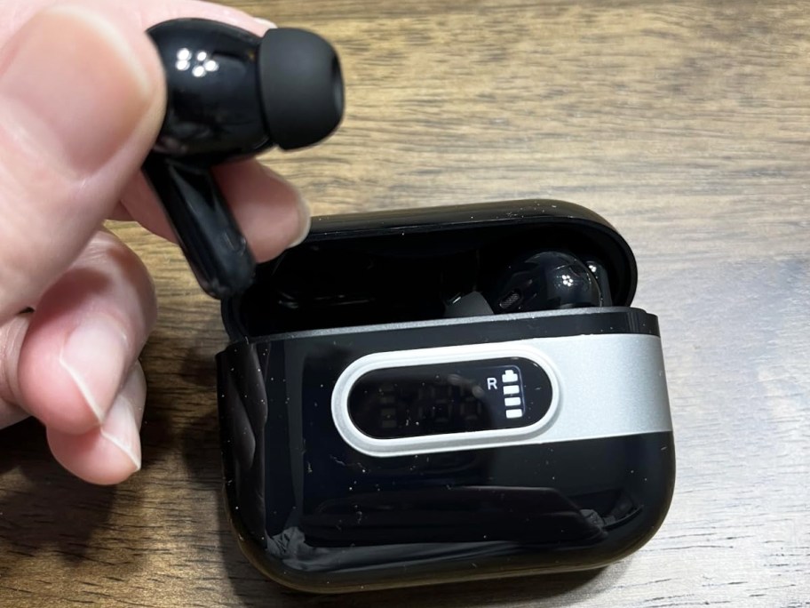 hand holding a black earbud with charging case behind it