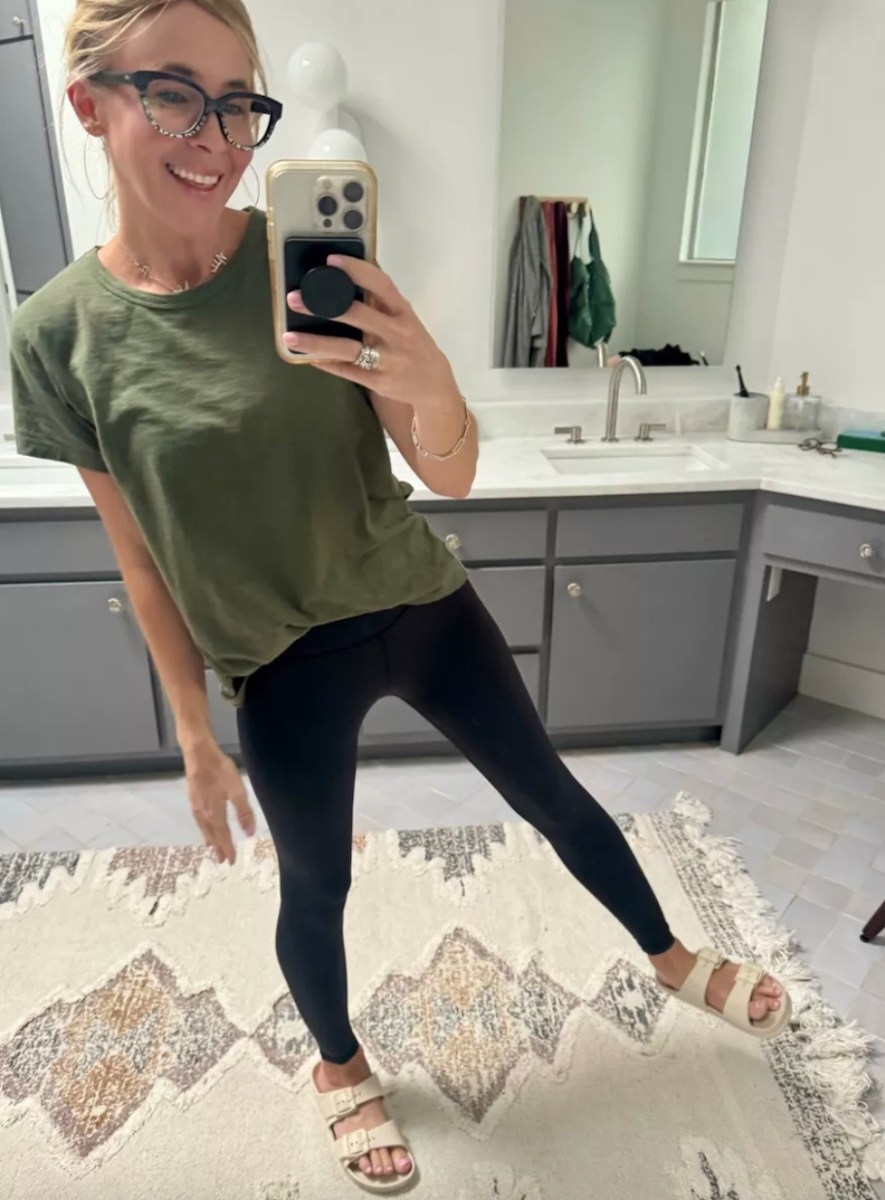 woman taking mirror selfie wearing green tee black leggings and slides