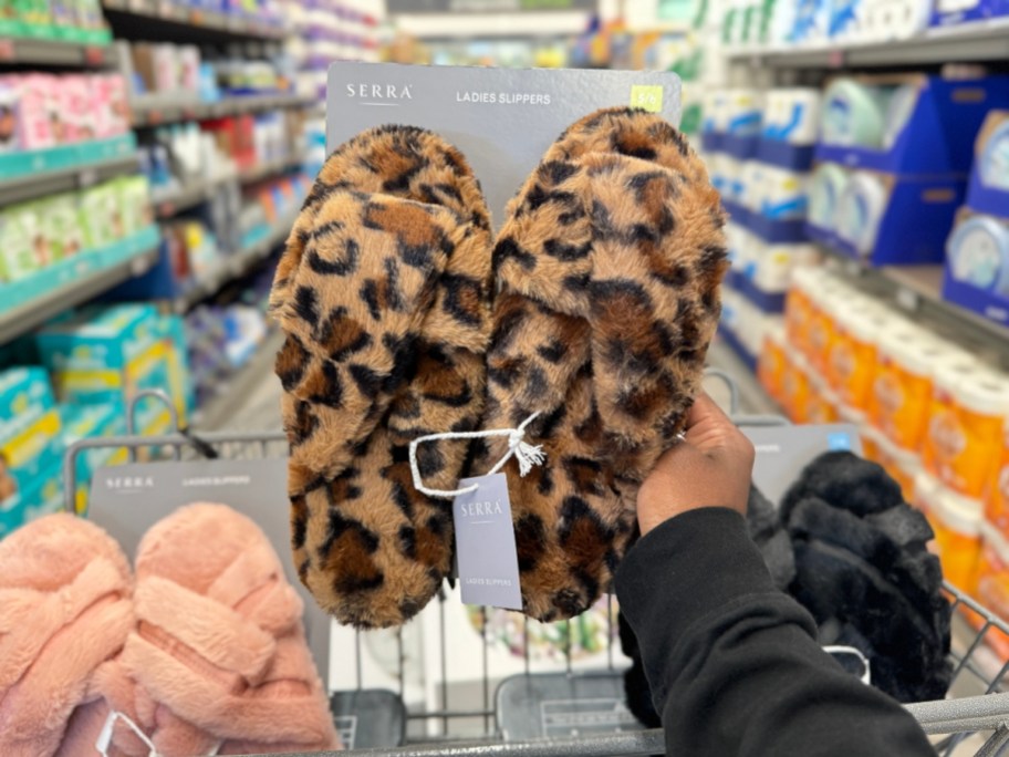 hand holding pair of leopard print fuzzy slippers, with a pink and black pair beside it